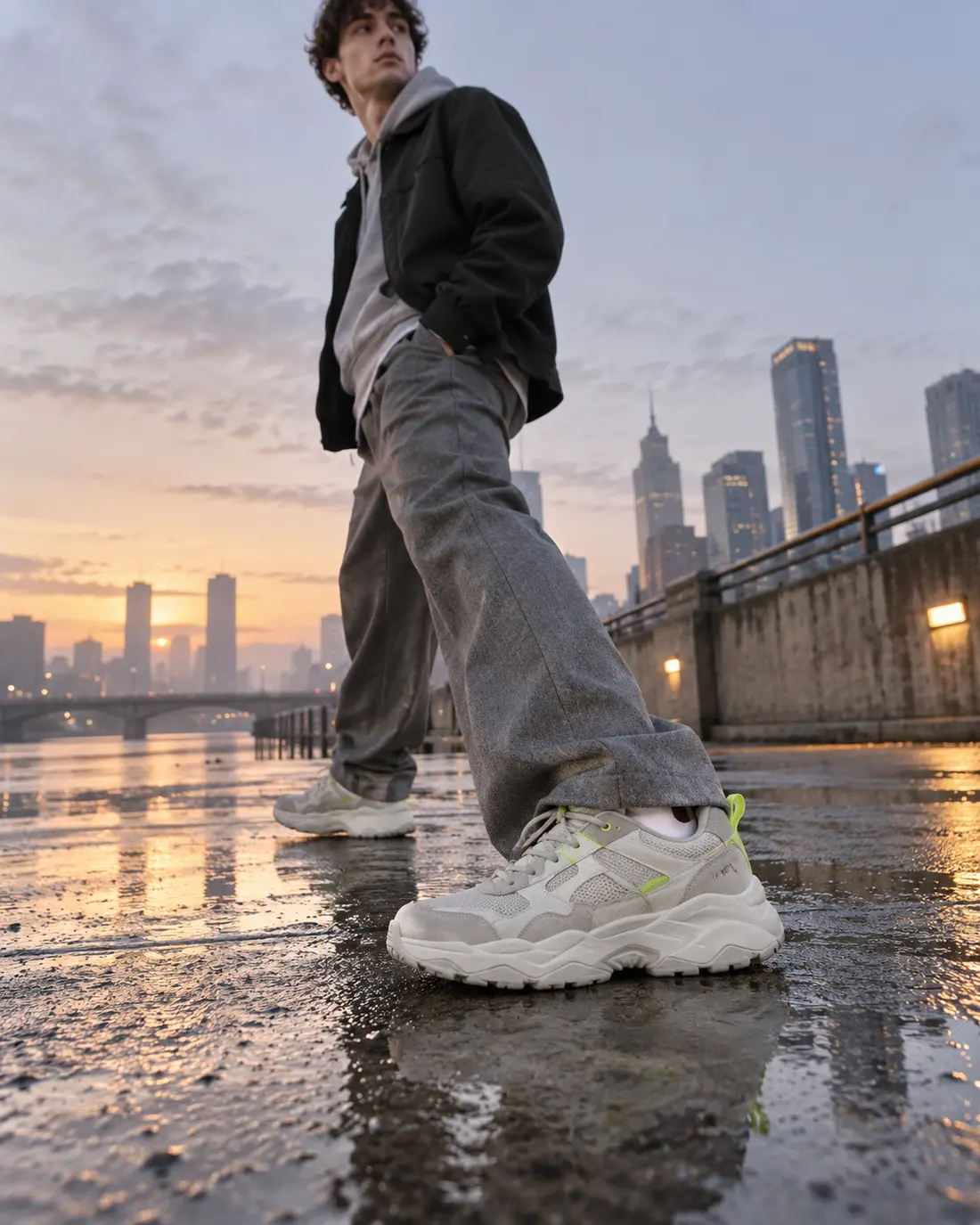 Man in wide grey trousers mid-stride on wet concrete at dawn in chunky off-white sneakers
