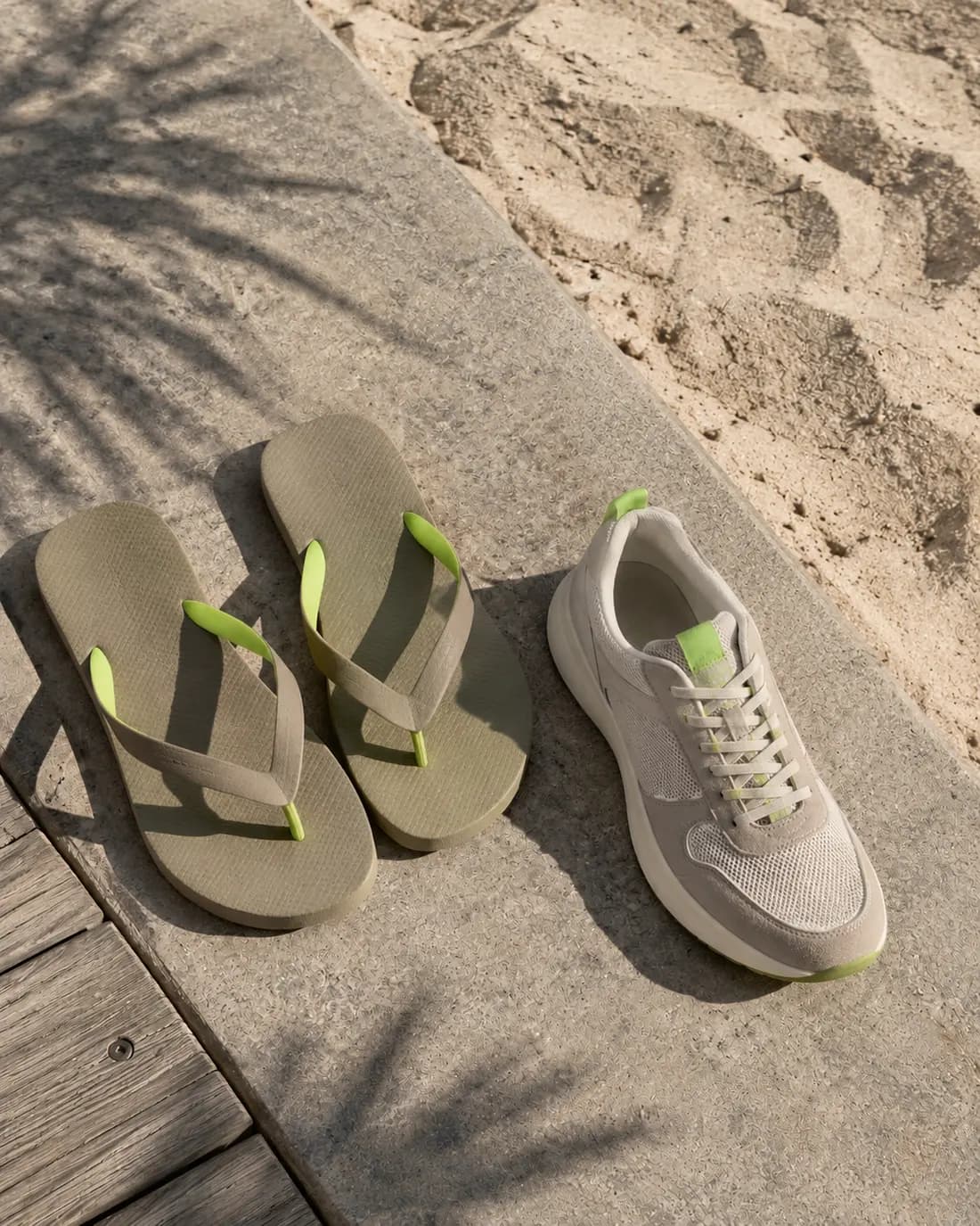 Flip-flops and a sneaker resting on a boardwalk where concrete meets sand